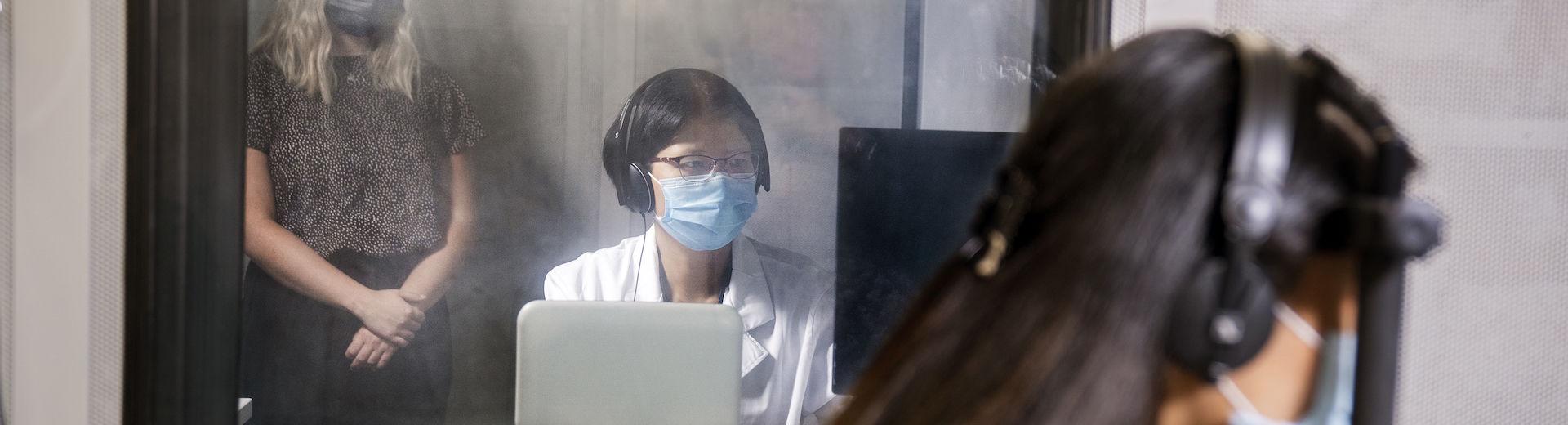 A masked student in a white shirt sits at a laptop with a woman standing behind here.